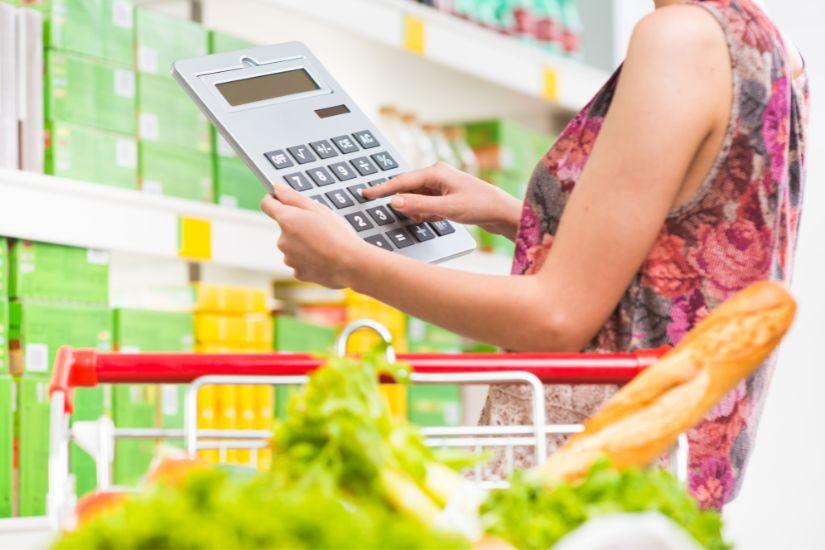 Es ist unbestritten, warum du dein Budget immer wieder anpassen musst, damit es optimal zu deinem Leben und Zielen passt. Eine Frau im Supermarkt beim Einkaufen mit einem großen Taschenrechner.